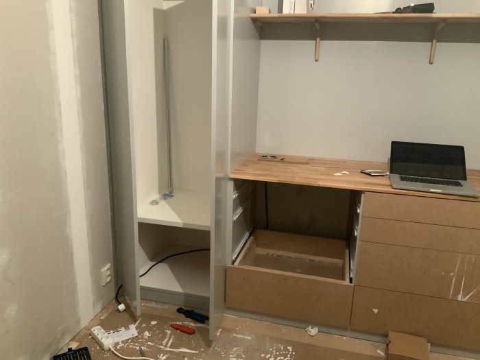 Interiör av ett pågående byggprojekt med osammanbyggd garderob och en arbetsbänk med öppen dosa och kabel.