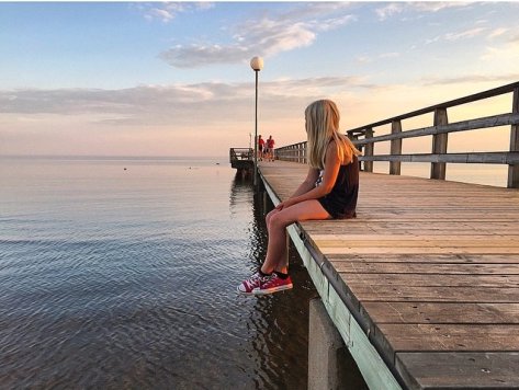 Person sitter på kanten av en träbrygga och tittar ut över havet vid solnedgång.