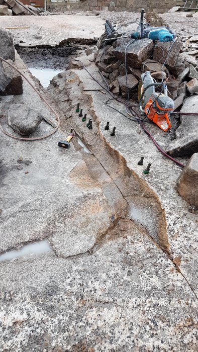 Granitberg med insatta kilar längs spricklinje, omgivet av arbetshandskar, kilverktyg och en motorkap.