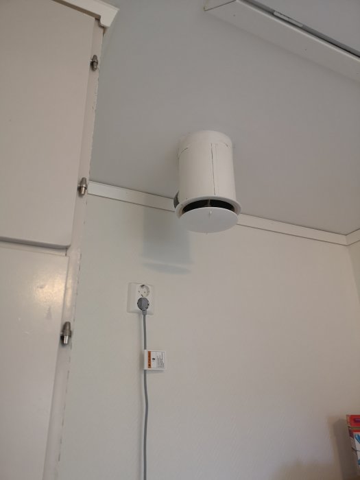 Vit Paxfläkt monterad i taket bredvid en elkontakt och ett stängt fönster i ett rum.