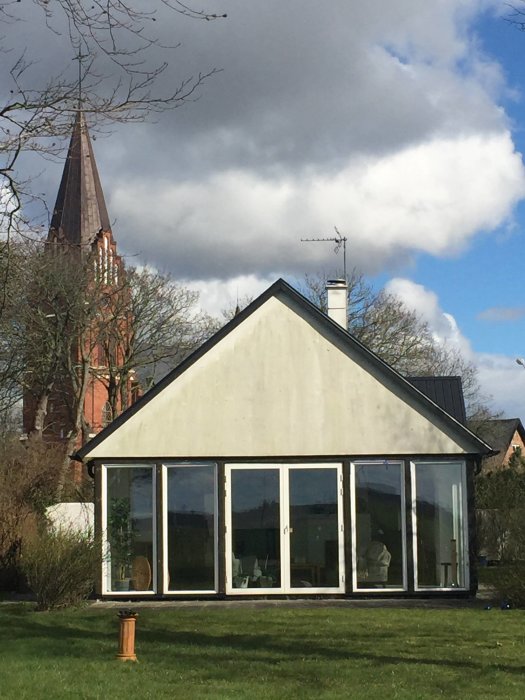 Uteplats med stora fönsterpartier på ett hus, gräsmatta framför och kyrktorn i bakgrunden.