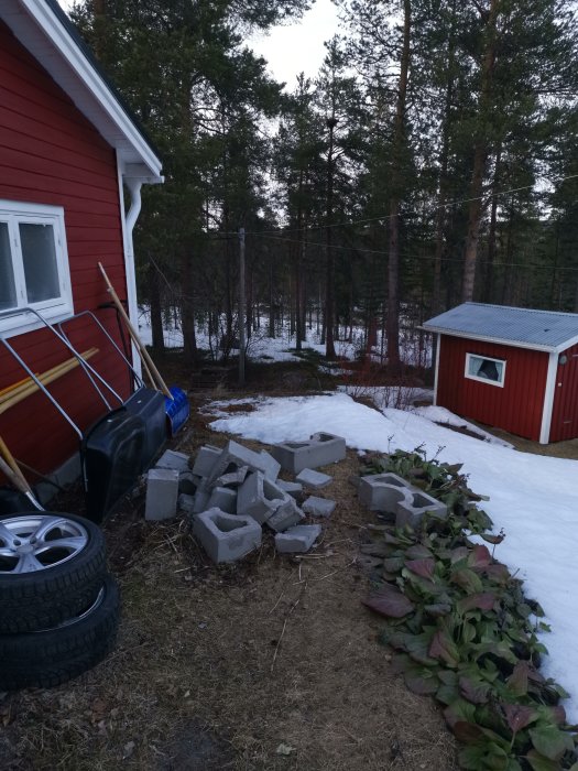 Hus med röda väggar och en hög med gråa tegelstenar på marken framför, omgivet av snö och träd.