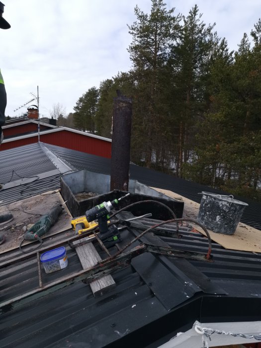 Rostig skorsten och verktyg på ett tak under renoveringsarbete med skogen i bakgrunden.