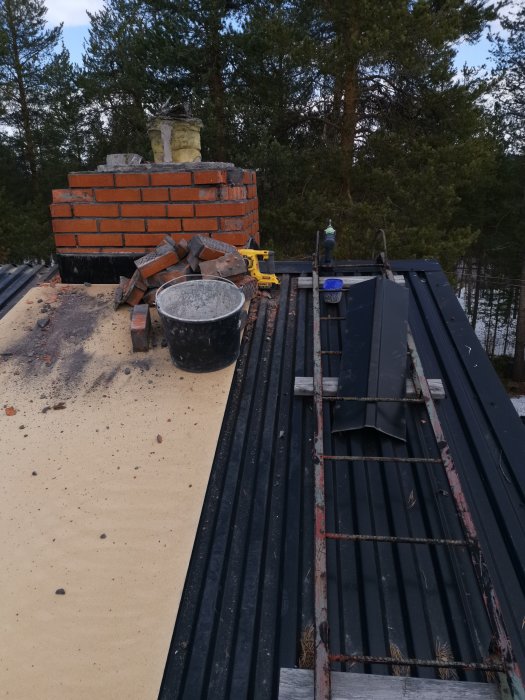 Nedmonterad skorsten med tegelstenar och verktyg på ett plåttak mot träd.