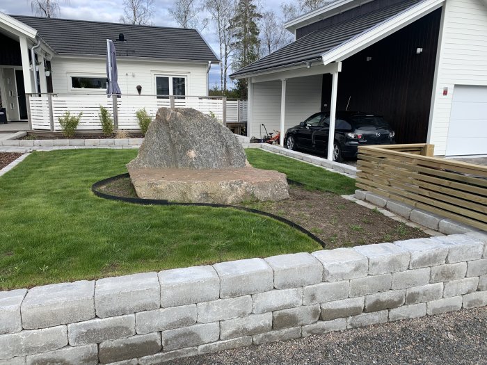 Grönt gräs omringar en stor sten i en trädgård framför ett vitmålat hus och garage med parkerad bil.