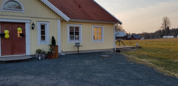 Gult hus med amerikansk veranda i påbyggnad, röda takpannor och omgiven av landskap.