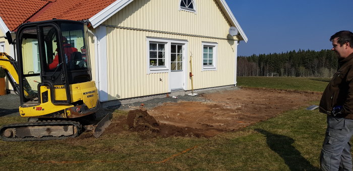 En grävmaskin framför ett gult hus under arbetet med grunden till en tillbyggnad av en veranda, med avgrävd mark och verktyg synliga.