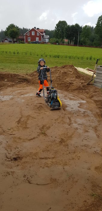 Barn i arbetskläder hanterar en markvibrator framför en hög med jord på byggarbetsplats.