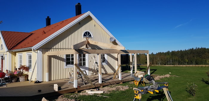 Utbyggnad av trähus med pågående konstruktion av veranda under blå himmel.