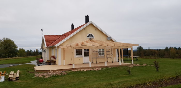 Gult hus med påbörjat tillbygge av träkonstruktion för veranda under molnig himmel.