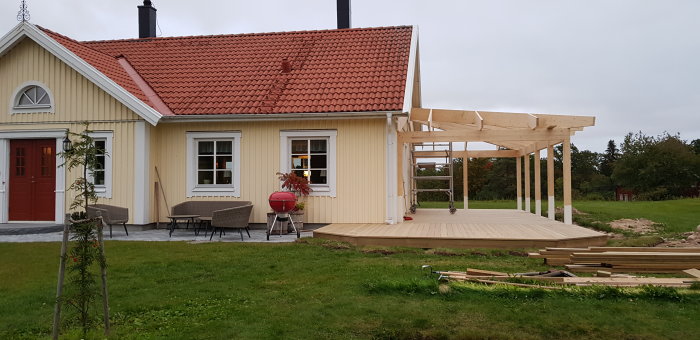 Ett gult hus med rött tegeltak där en ny träterrass och pergola är under konstruktion.