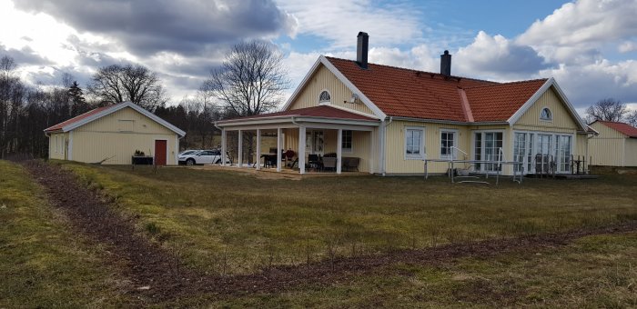 Ett gult hus med rött tak och en veranda, samt en fristående garagebyggnad och en bil parkerad utanför.