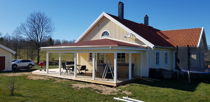 En nybyggd veranda med vitmålade pelare och räcke vid ett gult hus med rött tak under en klarblå himmel.