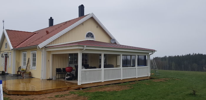 Gult hus med röd takpannor och nybyggd veranda under grå himmel.