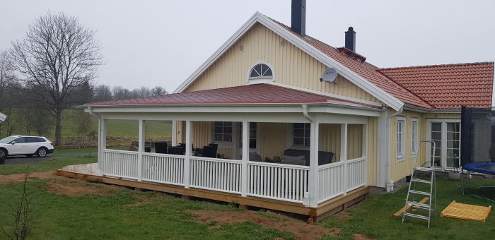 Nybyggd veranda med vit räcke på ett gult hus med röd takpannor och grå himmel i bakgrunden.