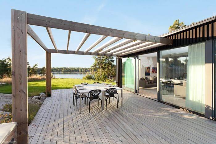 Modern pergola konstruerad av stora träbjälkar med utsikt över vattnet, angränsande till en trätrall och hus.