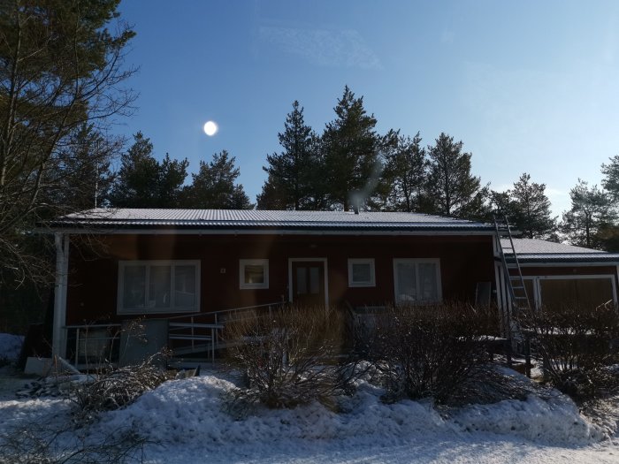 Ett enskilt hus täckt av ett tunt lager nysnö mot en bakgrund av soligt väder och tallar.