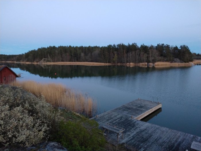 Kvällsutsikt över en lugn sjö med en brygga, ett rött skjul och skog i bakgrunden.