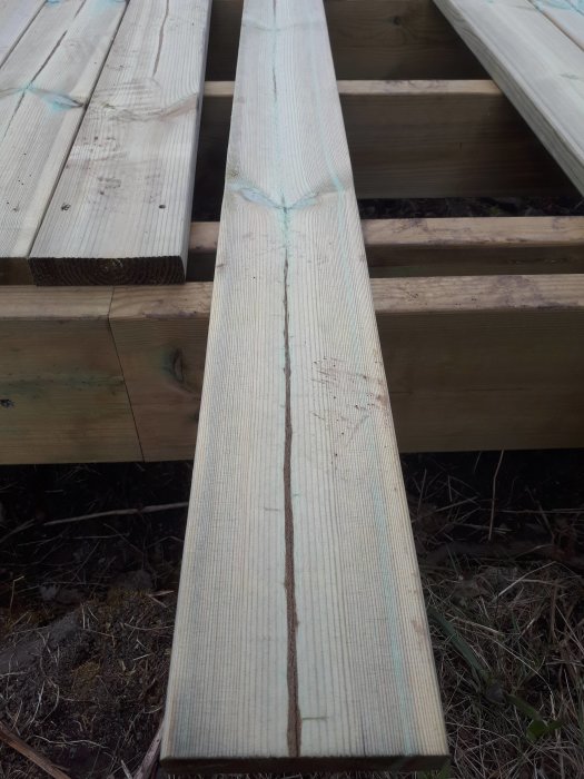 En träplanka med synlig kärna och bruna streck använd för trall, vilande på en träkonstruktion utomhus.