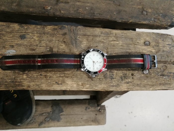 Armbandsur med randigt band på grovt träunderlag, rödsvart bezel, vit urtavla.