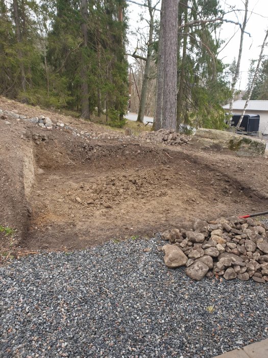 Uppgrävd plats i naturen för bygge av fristående förråd, omgiven av makadam och större stenar.