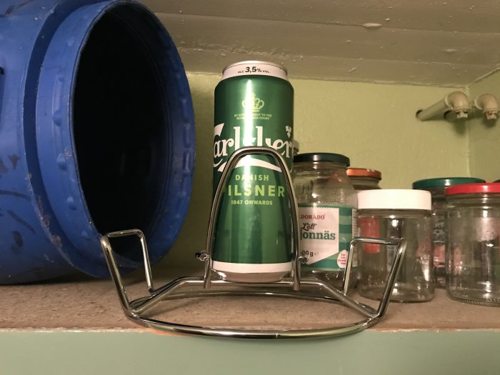 En 3,5:a Carlsberg ölburk står på en hylla bredvid ett antal glasburkar, framför en blå behållare och en metallställning.