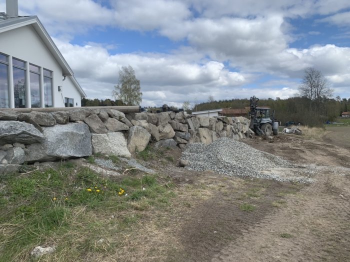 Nybyggd stenmur längs ett hus under klar himmel med en grävmaskin och grushög i bakgrunden.