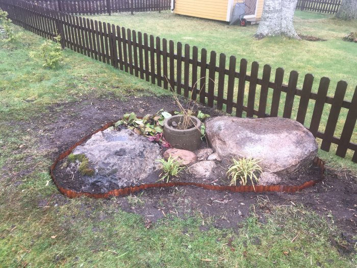 En nyplanterad rabatt med några halvvissna höga gräs, stora stenar och en komposterbar kruka vid ett staket.