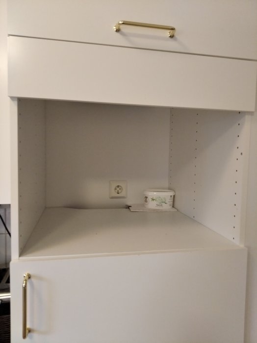 Öppet köksskåp med eluttag avsett för inbyggnadsugn, tomma hyllplan med hål för justerbara hyllor.