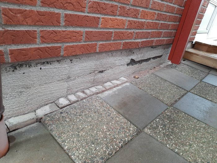Hålighet i putsen vid grunden av en tegelvägg nära ett hörn med röd dörr och stenplattor framför.