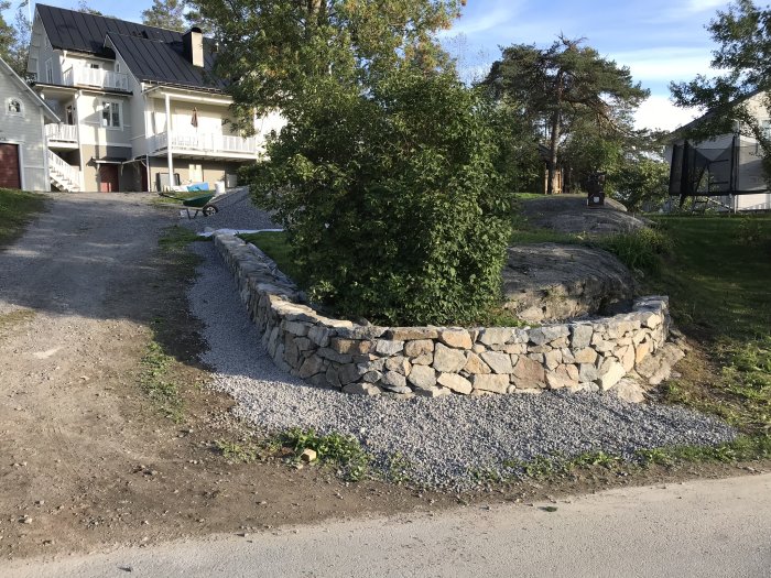 Två hemmabyggda stenmurar i trädgård med grönska och en vit villa i bakgrunden.