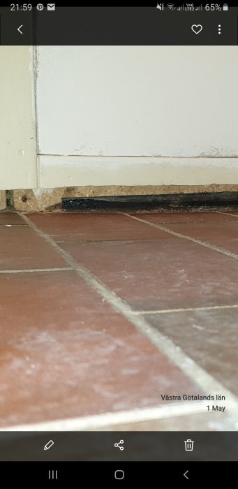 Golvsockel med en urjackad del som visar en svart, tjärliknande substans mot bakgrund av terrakottafärgade klinkerplattor.