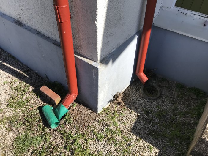 Husvägg med stuprör och en gammal dagvattenbrunn täckt av ett grönt rör och grus på marken.