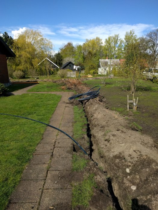 Grävt dike längs en trädgårdsgång med en slingrig vattenslang och grannhus i bakgrunden.