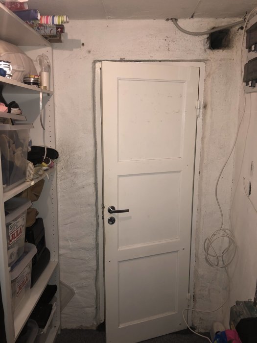Ojämna källarväggar med vit kalkfärg och stängd vit dörr, hyllor med föremål till vänster.
