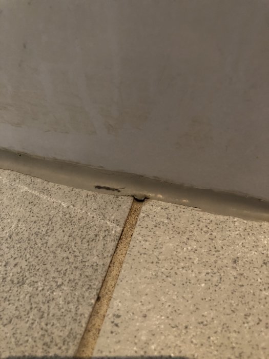 Slitna fogar mellan gråa klinkerplattor på golv och vita kakelplattor på vägg i ett badrum.