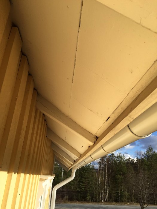 Takkanten på ett gult hus med möjliga eternitskivor under taköverhänget, nära en vit stupränna.