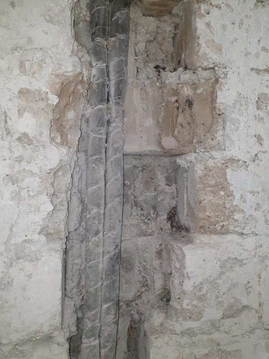 Nakna vattenrör i en ojämn källarvägg där gammalt kakel har tagits bort, med synligt puts och hålrum.