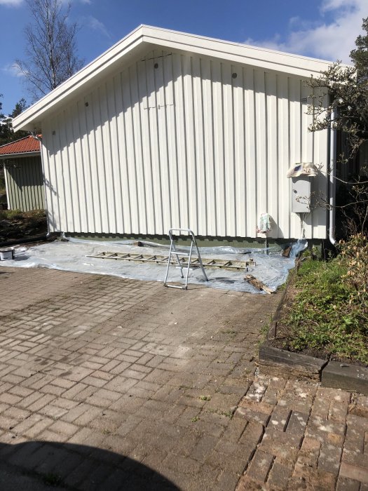 Ny målad vit fasad på hus med en bit av gammal färg synlig, stegpall och presenningar på marken.