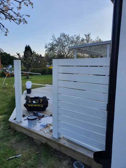 Hälften byggd vit altan med verktyg och byggmaterial ovanpå däcket, i en trädgård under konstruktion.