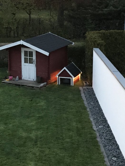 Trädgårdsbild på ett litet rött hus med vitt tak och mindre röd gräsklipparbod, båda med belysning, i skymningen.