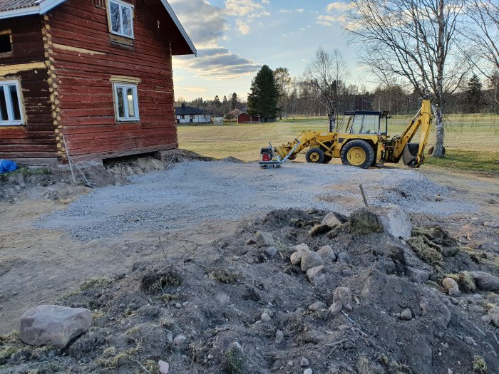 Markarbeten vid rödfärgad stuga, makadam utspridd framför, grävmaskin och paddningsmaskin syns.