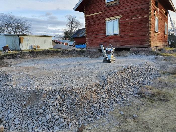 Markarbete med makadam och paddningsmaskin framför en byggnad i förberedelse för betonggjutning.