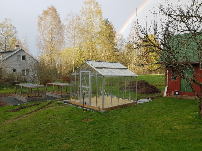 Växthus med nyligen installerade glaspaneler i en trädgård med regnbåge i bakgrunden.