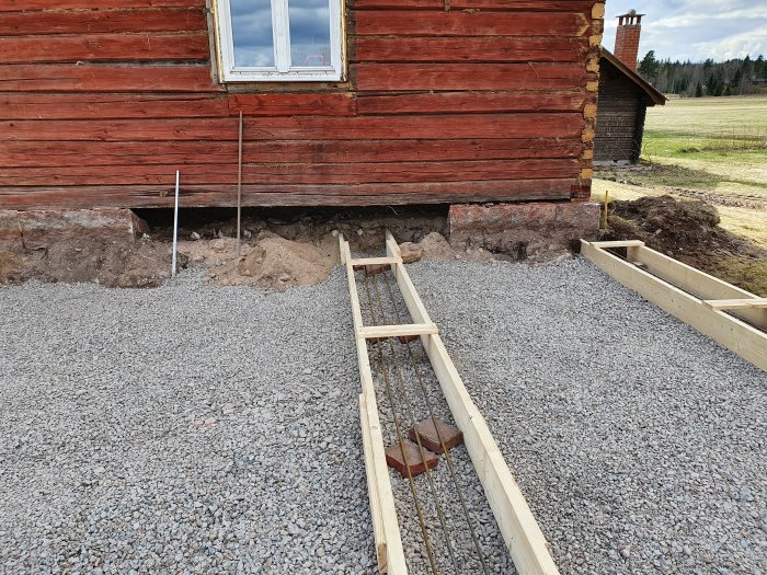 Pågående markarbete med makadam och trägjutform vid sidan av ett rött trähus.