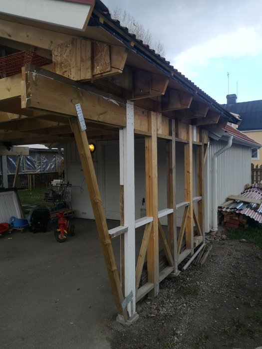 Halvfärdig carport med trästolpar och stödben, med oron att konstruktionen kan vika sig.