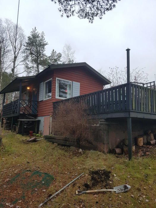 Rödmålad stuga med svart veranda och utskjutande tak över en mindre veranda, omgiven av grönska och verktyg på marken.