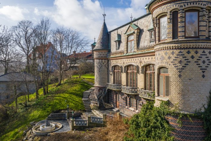 Ett storslaget slott med torn och utsmyckad fasad, ligger solbelyst omgivet av grönska och en fontän framför.