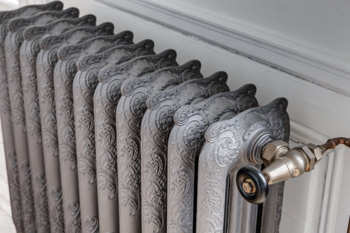 Ornately patterned vintage radiator with decorative swirls, connected to a brass valve, against a white wall.