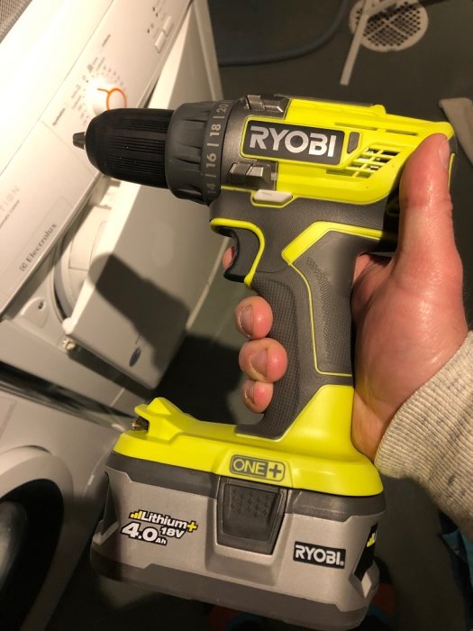 Hand håller en ny Ryobi borrskruvdragare med Lithium+ 4.0Ah batteri framför en vit tvättmaskin.